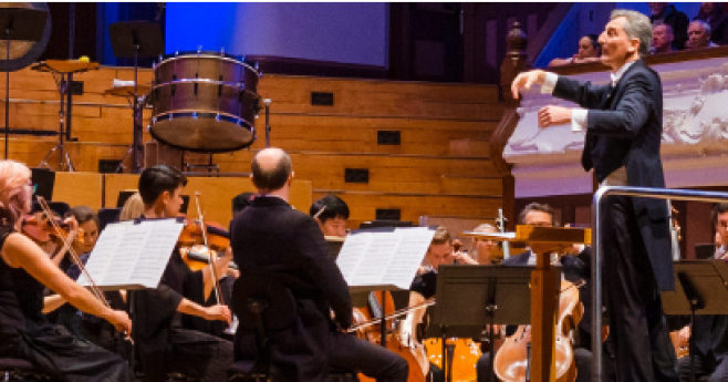 A conductor conducting the Auckland Philharmonia Orchestra in a large concert hall.